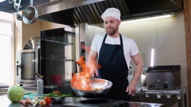 Şef mutfak restoranında ateşle yemek pişiriyor. Mutfakta tavayla yemek pişir. Üniformalı aşçı erkek, tavayı ateşle tutun. Kızarmış sebze pişiriyorum.