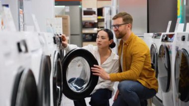 Genç çift ev aletleri mağazasında çamaşır makinesine bakıyor. Farklı çamaşır makineleri, seçim yap ve satın al. Evli bir çift, ev aletlerini hipermarketten seçer.