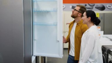 Hırdavatçıdaki genç çift ev için buzdolabı almayı seçiyor. Bir sürü buzdolabı var, seç ve al. Evli bir çift, ev aletlerini hipermarketten seçer.