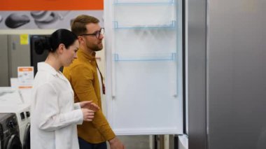 Donanım elektronik mağazasındaki buzdolabının kapısını açıp diğer modellerle karşılaştırın. Bir sürü buzdolabı var, seç ve al. Evli bir çift ev aletlerini seçer.