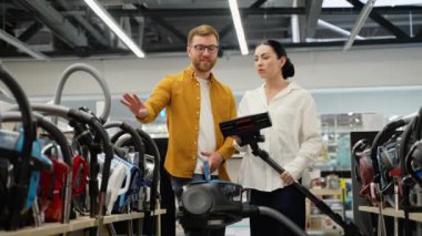 Genç bir çift elektrik süpürgesi seçerken tartışıyor. Yeni teknoloji konsepti. Alışveriş zamanı eğleniyor. Ev aletleri mağazasında genç bir çift.