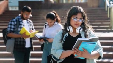 Elinde kitap tutan genç Hintli üniversiteli kız, arka planda okul binasına karşı bulanık bir grup arkadaşla kameraya poz veriyor. Gençlerin yakın arkadaş kavramının öğrenilmesi ve arkadaşlığı