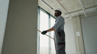 Afrikalı Amerikalı ressam evin duvarını boyuyor. Afrikalı Amerikalı müteahhit, inşaat alanının içindeki beton duvara astar beyaz boya yapmak için uzun saplı paten fırçası kullanıyor.