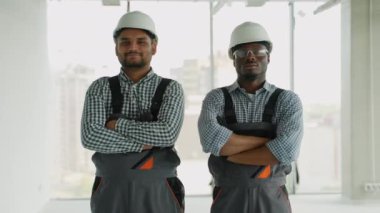 Hintli ve Afrikalı Amerikalı mühendis veya teknisyen kameraya gülümseyen ve kollarını kapalı inşaat alanında çaprazlayan bir şekilde bakıyor.