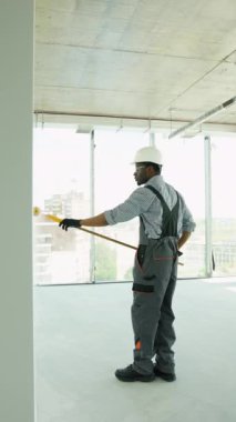 Afrikalı Amerikalı ressam ofiste bir duvar boyuyor. Afrikalı Amerikalı müteahhit, inşaat alanının içindeki beton duvara astar beyaz boya yapmak için uzun saplı paten fırçası kullanıyor.