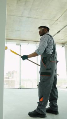Afrikalı Amerikalı ressam ofiste bir duvar boyuyor. Afrikalı Amerikalı müteahhit, inşaat alanının içindeki beton duvara astar beyaz boya yapmak için uzun saplı paten fırçası kullanıyor.
