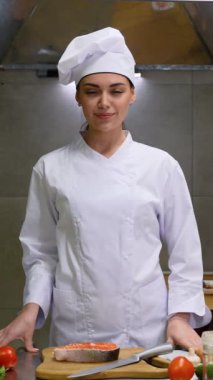 Otel mutfağında duran taze somonla gülümseyen beyaz kadın şefin portresi.