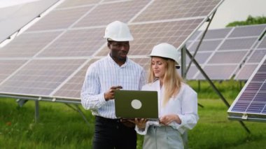 Dizüstü bilgisayar kullanan çeşitli kadın ve erkek mühendisler kırsal kesimde fotovoltaik pillerin yanında dururken projeyi tartışıyorlar.