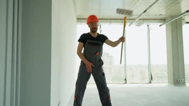 Komik inşaatçı inşaat alanında uzun kulplu fırçayla dans eder ve şarkı söyler. Profesyonel ressam ofiste bir duvar boyuyor.