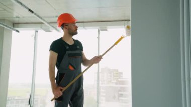 Ofiste duvar boyayan profesyonel bir ressam. İnşaat işçisi, inşaat alanının içindeki beton duvara astar beyaz boya yapmak için uzun saplı paten fırçası kullanıyor.