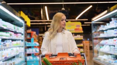 Alışveriş arabalı güzel sarışın kadın hareketli ve yoğun bir markette çeşitli ürünlere göz atıyor. Kafkasyalı kadın bir süpermarketin koridorlarında alışveriş yapıyor.