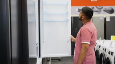 Alet gereçleri dükkanındaki buzdolabının kapısını açan Hintli adam yeni ev almak için diğer modellerle karşılaştırdı. Bir sürü buzdolabı var, seç ve al.