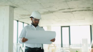 Hintli inşaat mühendisi bitmemiş bir binanın inşaat planlarını inceliyor. Güvenlik kaskı ve gözlük takıyor.