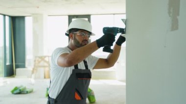 Hintli Asyalı inşaat işçisi beyaz bir şapka ve gözlük takıyor, bitmemiş bir binanın duvarına elektrikli matkap takıyor. Gri tulumlar giyiyordu ve...