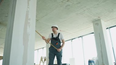 Bir inşaatçı, inşaat alanında bir silindir kullanarak duvara pürüzsüz bir kaplama uyguluyor. Güvenlik kıyafeti giyiyor.
