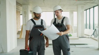 Hintli ve Kafkasyalı bir müteahhit bir inşaat alanının planlarını inceliyor. Her ikisinde de güvenlik donanımı var.