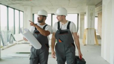 Hintli ve Kafkasyalı bir müteahhit, planları gözden geçirirken bir inşaat sahasında birlikte yürüyorlar. Her ikisinde de güvenlik donanımı var.