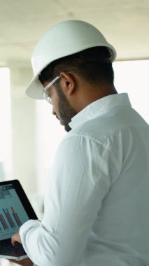 Beyaz gömlekli ve kasklı Hintli mühendis inşaat alanındaki bir dizüstü bilgisayardaki verileri analiz ediyor. Profesyoneller teknolojiyi projede hassasiyet ve verimliliği sağlamak için kullanıyorlar.