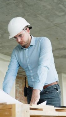 İnşaat mühendisi bitmemiş bir inşaat alanının planlarını dikkatlice inceliyor. Kask ve güvenlik gözlüğü takıyor.