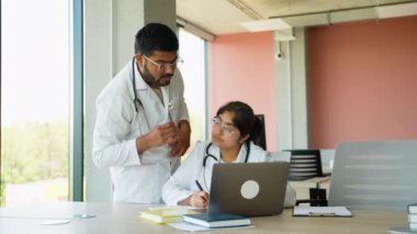 Beyaz önlüklü iki Hintli stajyer stajyer stajları sırasında ders çalışıyor. Bir dizüstü bilgisayar ve tıp kitapları kullanıyorlar, klinik kavramları tartışıyorlar ve modern bir çalışmada pratik görevler için hazırlanıyorlar.