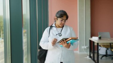 Beyaz önlük ve steteskop giyen Hintli tıp öğrencisi sınavlara hazırlanırken bir kitap okuyor. Üniversitede bir pencerenin yanında duruyor, tamamen derslerine ve tıp kariyerine odaklanmış.