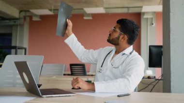 Hintli doktor, tıbbi teşhis koymak için röntgen görüntüsünü dikkatlice inceler. Steteskop takan ve klinik bir ortamda oturan doktor, hasta sağlığını değerlendirmeye odaklanmış durumda.