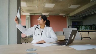Pakistanlı kadın doktor masasında oturmuş teşhis üzerinde çalışırken röntgeni dikkatlice inceliyor. Profesyonel sağlık hizmetleri için modern ofis ortamında dizüstü bilgisayar ve tıbbi notlar kullanıyor.