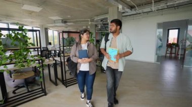 Two Indian students are walking down the university corridor with books and backpacks, engaged in conversation. The video portrays student life, education, and collaboration in a modern academic