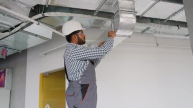 Hintli teknisyen bir ofisteki havalandırma sistemini tamir etmeye çalışıyor. Görüntü, modern binalarda etkin hava dolaşımı için bakımın önemini vurguluyor.