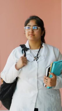 Hintli tıp öğrencisi elinde bir sırt çantası ve elinde kitaplarla üniversite koridorunda yürüyor. Beyaz önlük ve steteskop takarak tıp eğitimine ve geleceğine bağlılığını temsil ediyor.