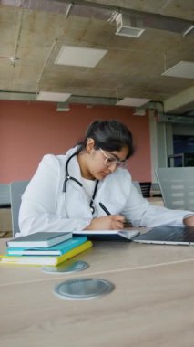 Yorgun bir Hintli kadın doktor, klinikteki stresli bir günün ardından yorgunluktan başını tutuyor. Etrafı iş malzemeleriyle çevrili, sakin ofis ortamındaki zorlu günü yansıtıyor.