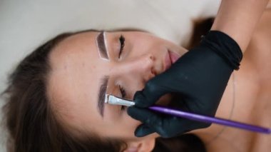 Bir güzellik teknisyeni kalıcı makyaj uygulaması için bir fırça kullanarak kaş hatlarını dikkatlice tanımlar. Kaşları kusursuz bir görünüş için hazırlanırken müşteri sakin kalıyor.