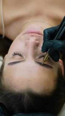 Kalıcı kaş makyajı uygulamasının yakın çekimi. Teknisyen, dövme makinesi kullanarak hassas bir şekilde çalışıyor. Müşteri rahatlarken doğal ve uzun süreli bir sonuç elde ediyor.
