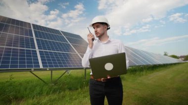 Avrupalı mühendis, bir güneş çiftliğindeki dizüstü bilgisayarındaki verileri analiz ederken telsizle iletişim kuruyor. Güneş panellerinin optimum verimliliğini sağlayarak enerji üretimini ve performansını izliyor.