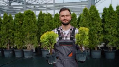 Üniformalı bir bahçıvan, müşteri için özenle seçilmiş iki saksı süs ağacı tutar. Etrafı çeşitli yeşil bitkilerle çevrili bir bahçe merkezinde duruyor.