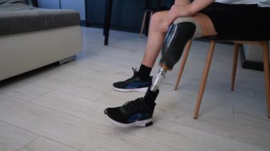 Modern dairesinde bir sandalyede oturan, protez bacağını ve spor ayakkabılarını gösteren bir adamın yakın plan fotoğrafı.