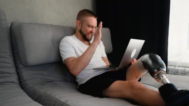 Protez bacaklı neşeli bir adam rehabilitasyon seansı için doktoruyla çevrimiçi video görüşmesi yapıyor. Video, teknolojiyle pozitiflik, esneklik ve modern sağlık hizmetlerini yakalıyor.