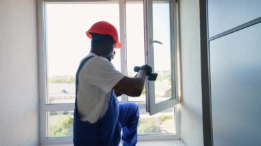 Mavi tulumlu ve başlıklı odaklanmış bir işçi elektrikli matkapla PVC plastik pencere çerçevesi kuruyor. Uygun kurulumu sağlamak için çerçeveyi dikkatlice ayarlar.