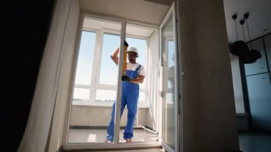 Mavi tulumlu ve kasklı yetenekli teknisyen, PVC pencere kurulumu sırasında kalite ve hassasiyete odaklanarak doğru hizalamayı sağlamak için bir seviye kullanır.