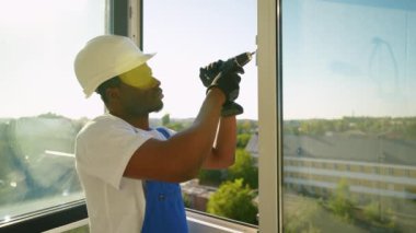 Güvenlik teçhizatında yetenekli bir teknisyen hassas bir noktaya odaklanmak için elektrikli matkap kullanarak PVC penceresi kuruyor. Parlak güneş ışığı pencereden sahneyi aydınlatır.