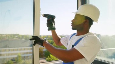 Güvenlik teçhizatında yetenekli bir teknisyen hassas bir noktaya odaklanmak için elektrikli matkap kullanarak PVC penceresi kuruyor. Parlak güneş ışığı pencereden sahneyi aydınlatır.