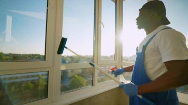 Mavi tulumlu ve eldivenli Afro-Amerikalı bir adam balkon camına sprey sıkar ve yıkamaya hazırlanır. Güneş ışığı işini aydınlatıyor, sıcak bir dokunuş ekliyor.