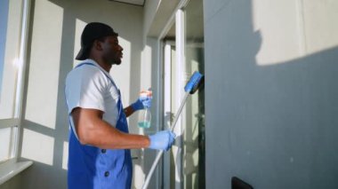 Mavi tulumlu profesyonel servis teknisyeni sprey ve cam sileceği kullanarak pencereyi temizliyor. Günışığı, modern bir dairede kusursuz çalışmalarını aydınlatıyor.