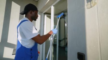 Kendini işine adamış mavi tulumlu ve eldivenli bir servis teknisyeni bir dairede cam sileceği ile pencereyi temizliyor. Güneş ışığı onun kusursuz temizlik çalışmalarını vurgular.