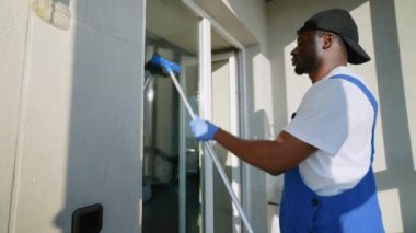 Kendini işine adamış mavi tulumlu ve eldivenli bir servis teknisyeni bir dairede cam sileceği ile pencereyi temizliyor. Güneş ışığı onun kusursuz temizlik çalışmalarını vurgular.