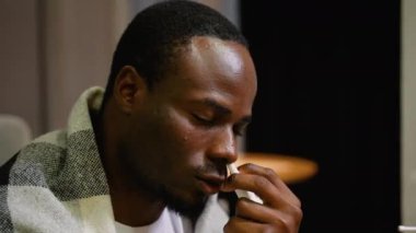 Tıkanıklık giderici olarak burun spreyi kullanan bir adamın yakın çekimi. Battaniyeye sarılmış halde, sıcak bir iç mekanda soğuk semptomların tesellisini arıyor.