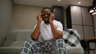 Bir adam kanepeye oturur, şakaklarını acı bir ifadeyle bastırır, gözle görülür bir şekilde şiddetli bir baş ağrısıyla mücadele eder. Vücut dili rahatsızlık ve gerilim gösteriyor.