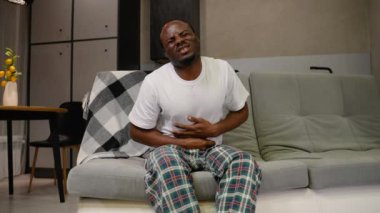 Afro-Amerikalı bir adam kanepeye oturmuş, acı dolu bir ifadeyle karnını sıkıyor, muhtemelen gıda zehirlenmesi ya da sindirim sorunları yaşıyor, görünür bir rahatsızlığı var.