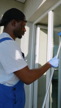 Mavi tulumlu bir temizlikçi, bir apartmanın pencerelerini temizlemek için sprey ve cam sileceği kullanıyor.