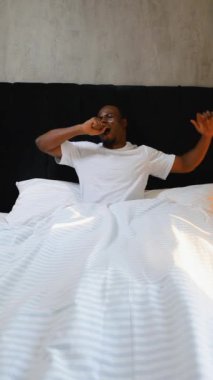 Afro-Amerikalı bir adam, sıcak sabah güneşinde yatağında uzanırken ve esnerken görülüyor. Minimalist yatak odası atmosferi sakin ve rahat bir hava katıyor, sakin bir anı yakalıyor.